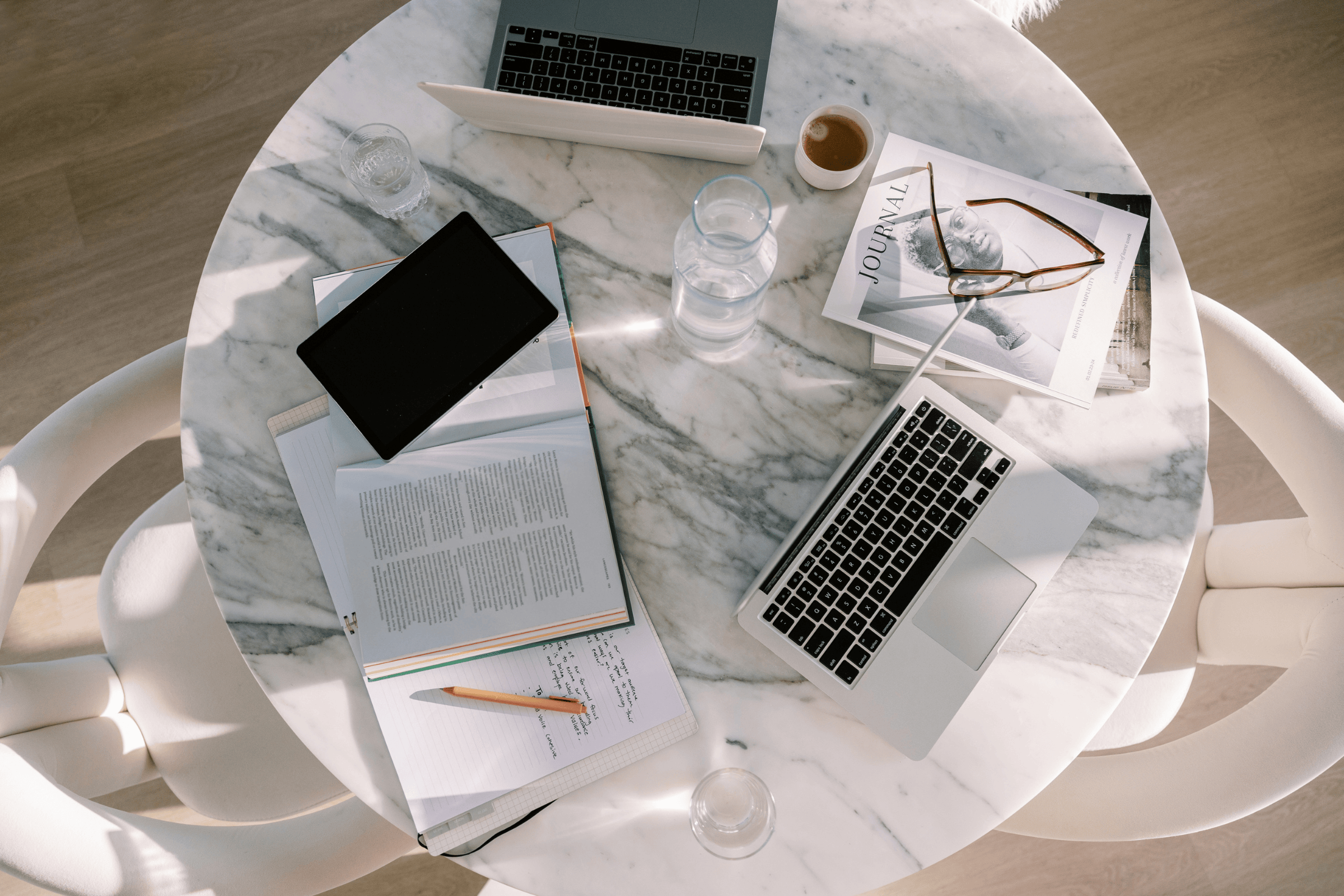 Overhead view of a marble desk in natural light with an open notebook of handwritten ideas, a pen, an open book, two laptops, and a coffee